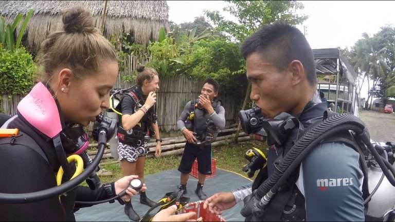 Two divers practising scuba skills on a training platform, with MCP's thatched-roof building behind them