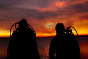 Silhouettes of two divers watching a dramatic orange sunset over the water