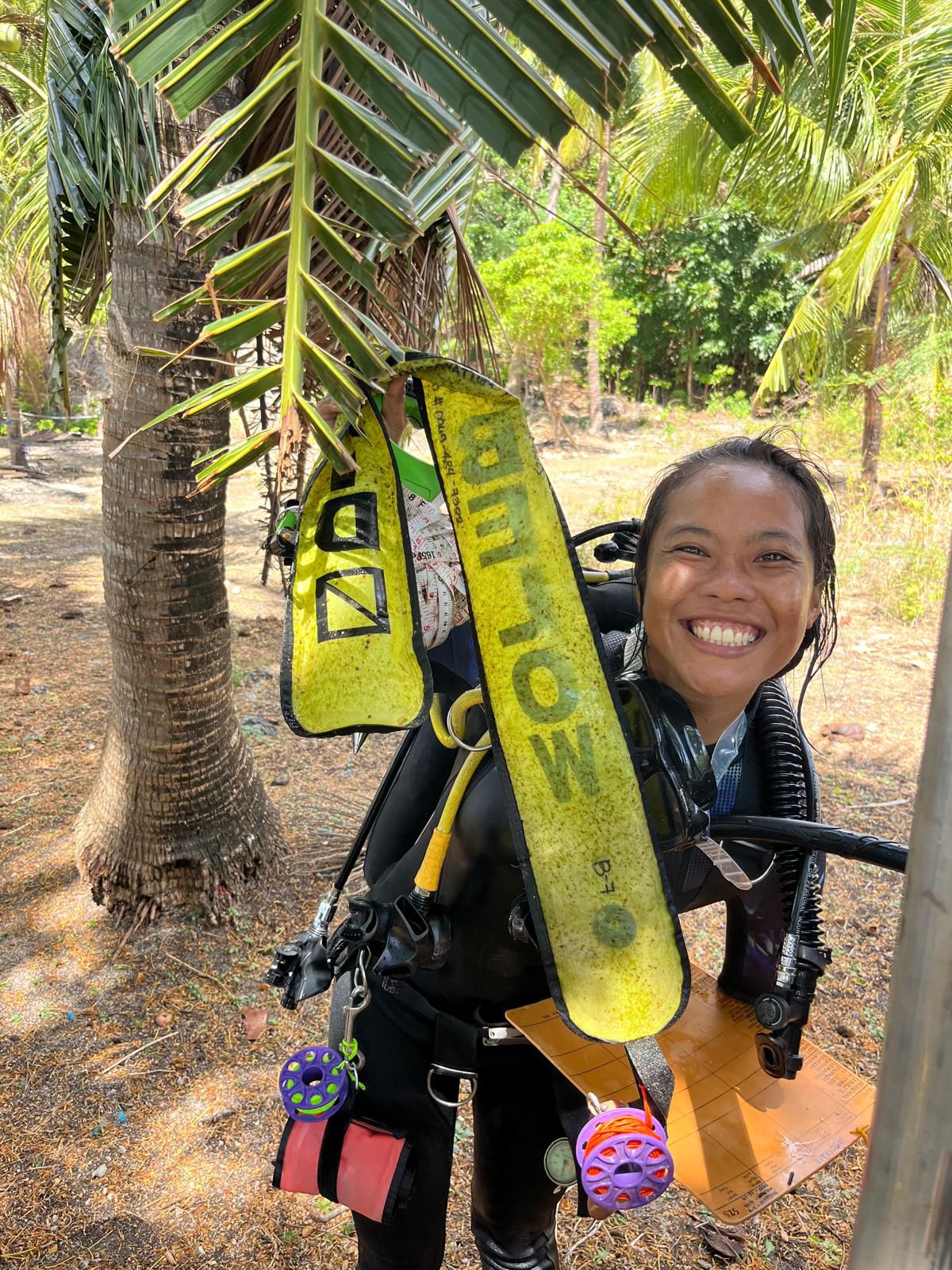 A scholarship student smiling in scuba gear among palm trees