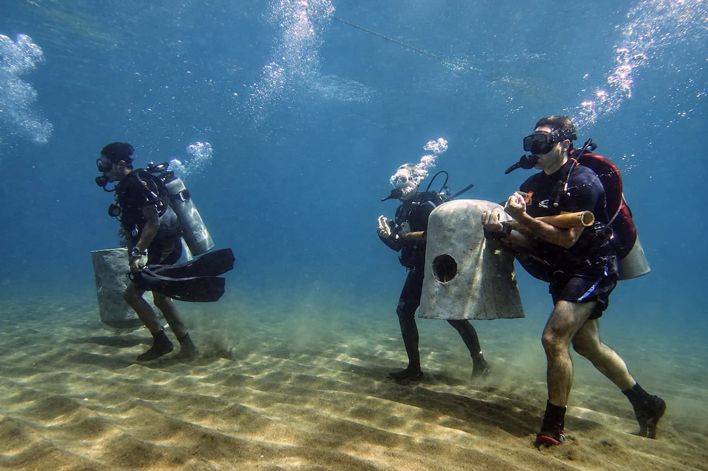 Divers carrying concrete reef modules across a sandy seabed to build an artificial reef
