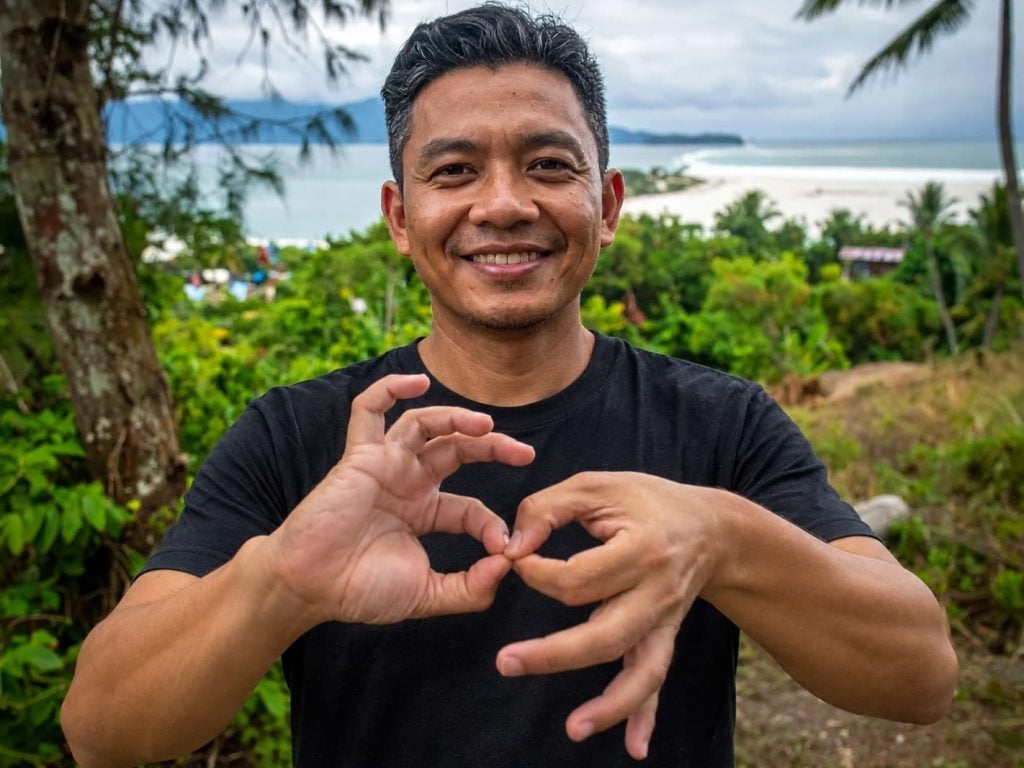 Smiling man making a hand sign, with the forest and coastline of southern Negros behind him