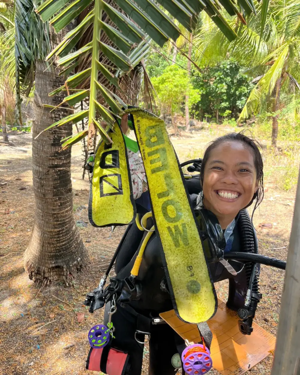 An MCP scholar smiling in full scuba gear among palm trees, holding her fins